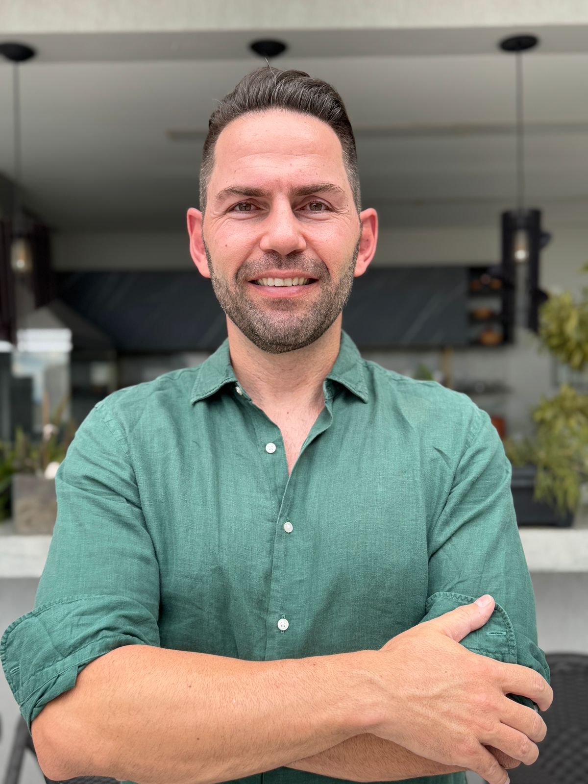 Email marketing agency founder in a green button-up shirt smiling confidently with arms crossed, standing in a modern indoor setting.